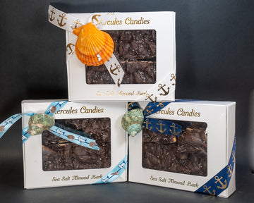 Three boxes of Hercule's Candies sea salt almond bark with decorative ribbons and shell accents on a dark background.